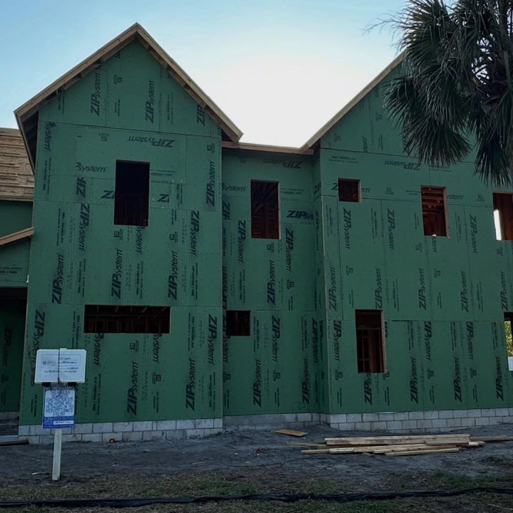 New residential home under construction with green sheathing, multiple windows, and a gabled roof, set against a clear sky and palm trees in the background.