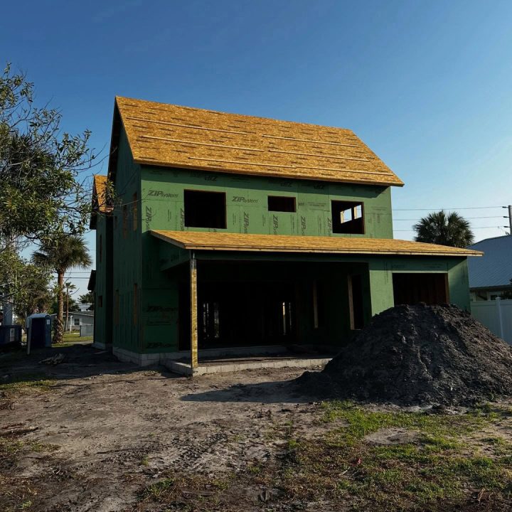 New two-story house under construction with green sheathing and a wooden roof, set against a clear blue sky, with a pile of dirt in the foreground.
