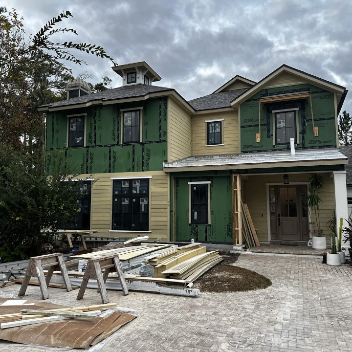 A detailed view of a new house under construction with green sheathing and beige siding, featuring multiple windows and a front porch area, set against a cloudy sky.