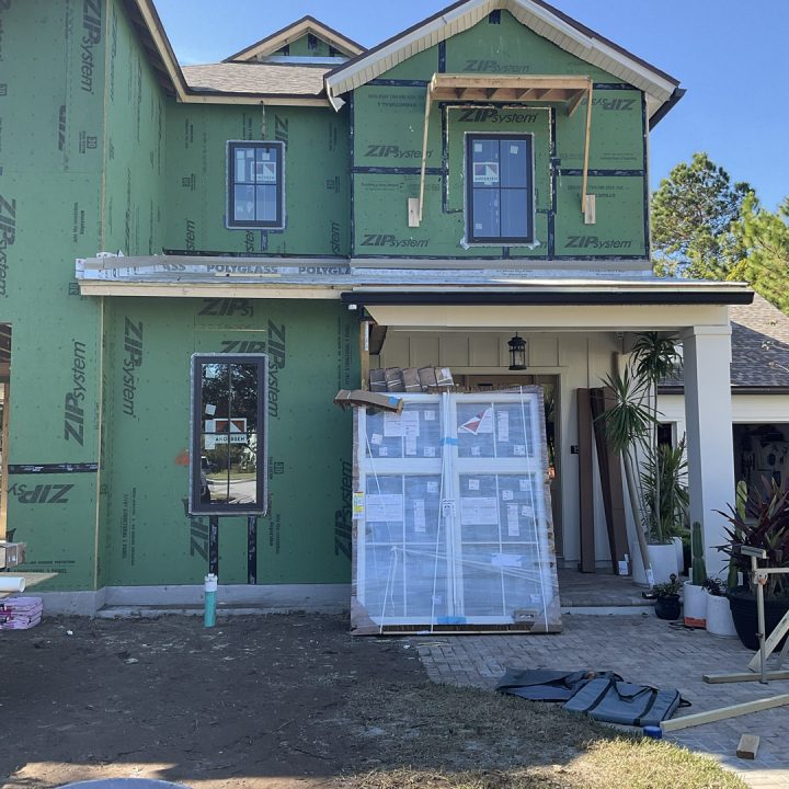 New two-story house under construction with green sheathing, windows, and front door installed, set against a clear blue sky.