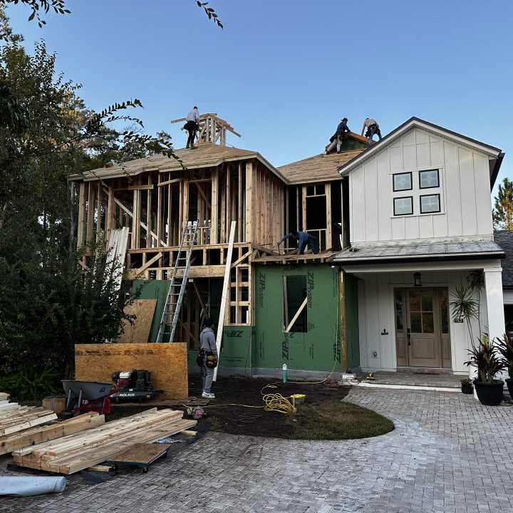 An outdoor view of a two-story house under construction, with workers installing the roof and framing the upper level, surrounded by trees and construction materials.