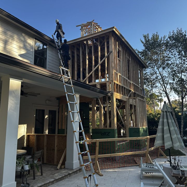 Construction workers building a second-story wooden deck on a residential home with a ladder and scaffolding, under a clear blue sky.
