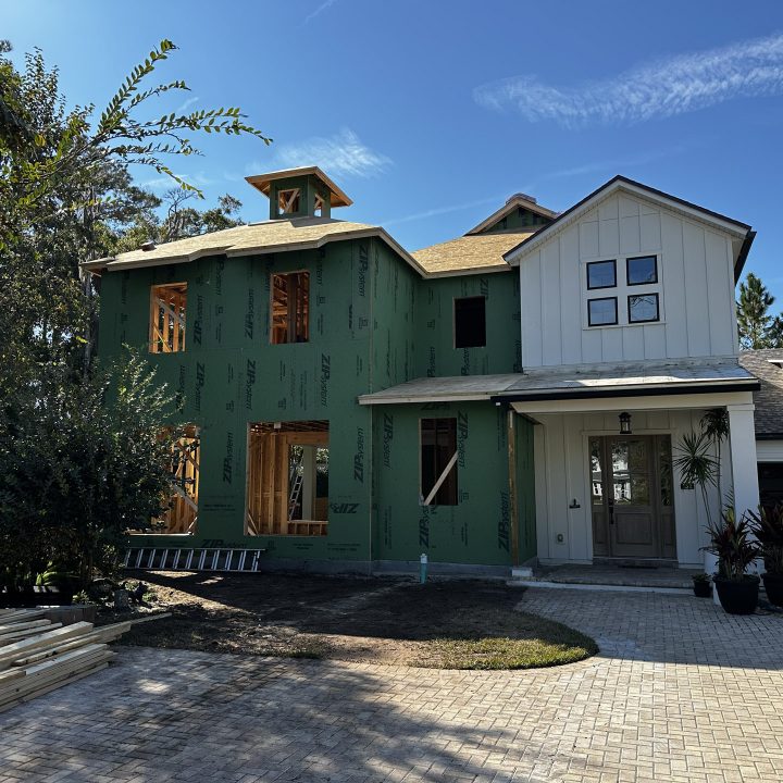 A detailed house under construction with a mix of green sheathing and finished white siding, featuring multiple windows, a garage, and a landscaped driveway, set against a bright blue sky.