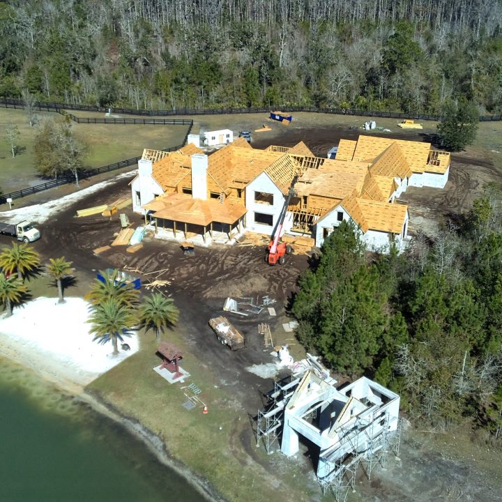 A construction site of a large, modern lakeside home in progress, with wooden framing and roofing being installed, surrounded by trees and a small dock on the water.