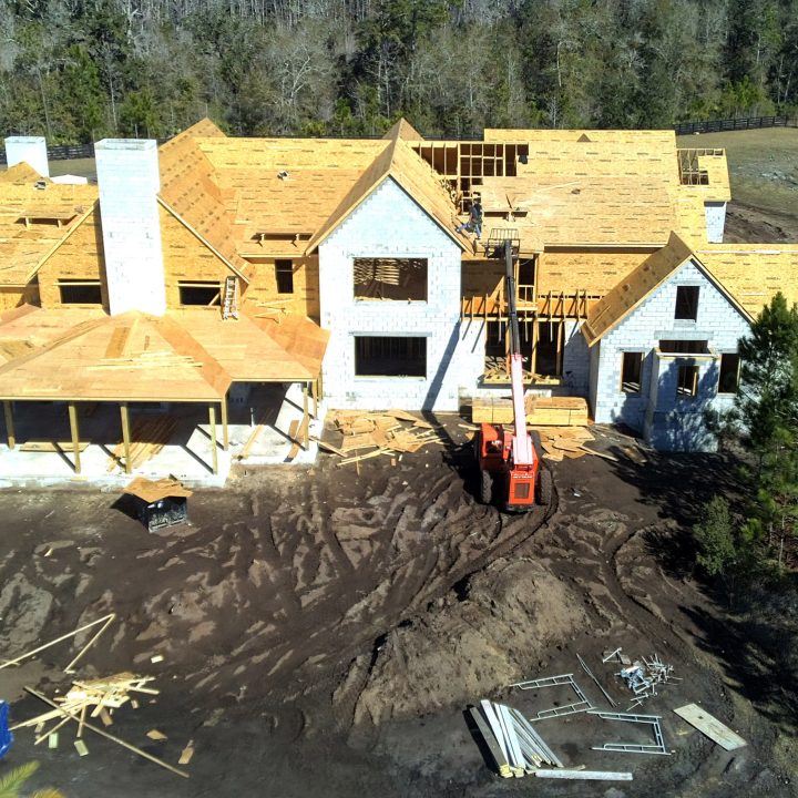 A detailed aerial view of a large, multi-story residential home under construction, with wooden framing and roofing in progress, surrounded by trees and construction equipment.
