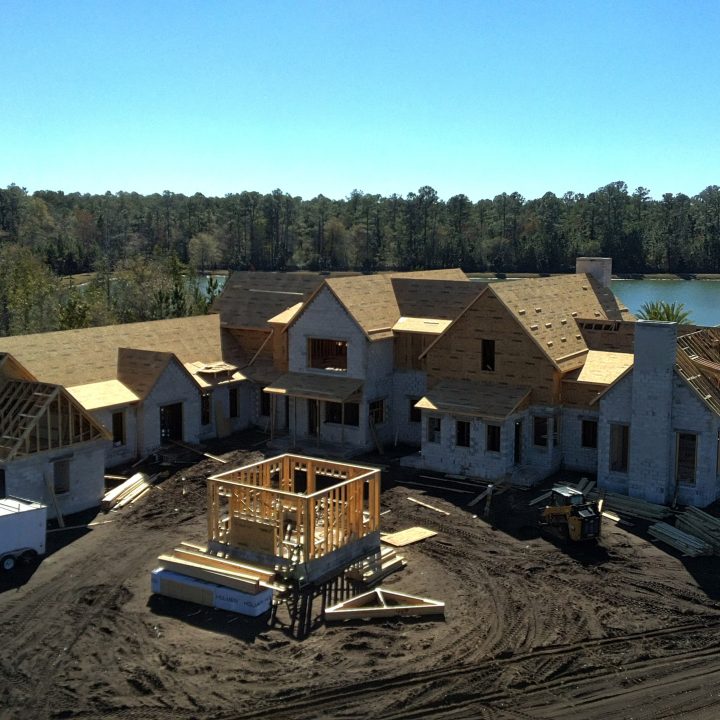 Modern house under construction with wooden framing and partially completed roof, situated near a water body with a clear blue sky overhead.