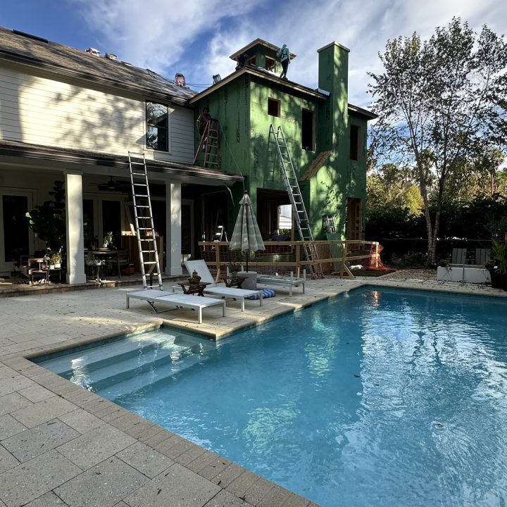 A detailed view of a house under construction with a green exterior, ladders, and scaffolding, situated next to a swimming pool with lounge chairs, under a partly cloudy sky.