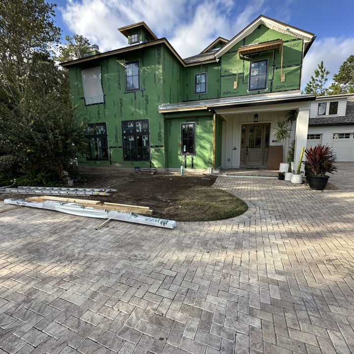 Newly constructed house with green exterior siding, multiple windows, and a front porch, situated on a paved driveway with landscaping and trees in the background.