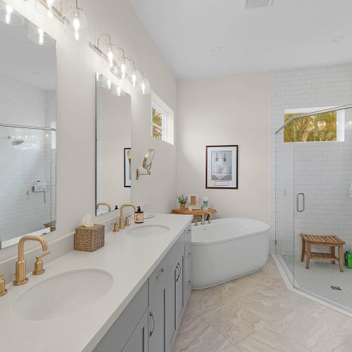 Bright and spacious bathroom featuring a double vanity with gold fixtures, a large mirror, a soaking tub, and a glass-enclosed walk-in shower with a wooden bench.