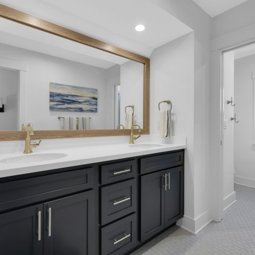 Spacious bathroom featuring a double vanity with dark cabinetry, two gold faucets, and a large wall-mounted mirror, illuminated by recessed lighting, with a window providing natural light.