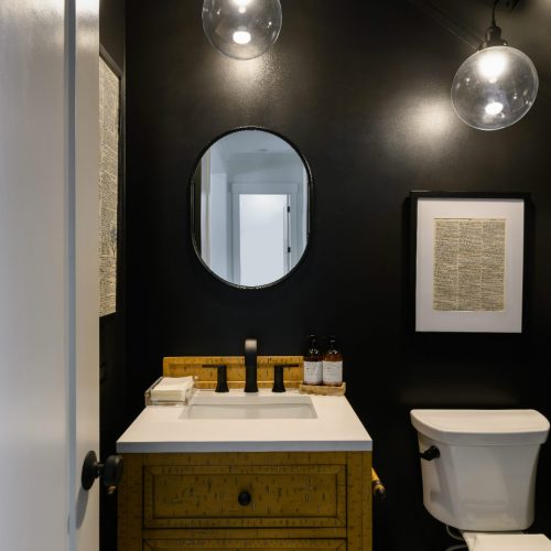 A contemporary bathroom featuring black walls, a wooden vanity with a white countertop, a round mirror, and modern lighting fixtures, creating a sleek and elegant space.