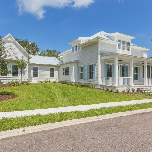 A white, two-story house with a wraparound porch, large windows, and a well-maintained lawn, showcasing a contemporary residential design by John Merrill Homes.