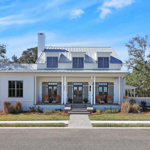 Beautiful white farmhouse with a spacious front porch, three front doors, and large windows, set against a clear blue sky with surrounding trees.