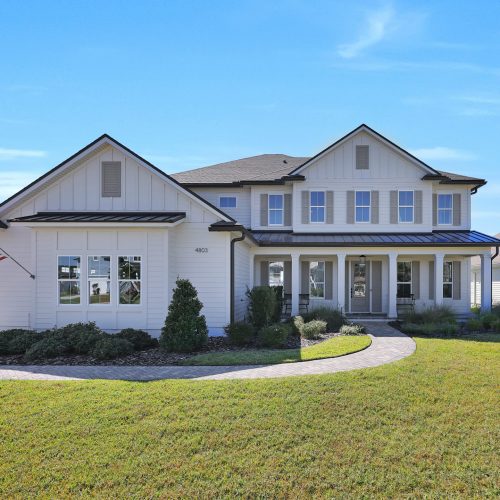 A spacious, newly built Craftsman-style house featuring a white exterior, large front porch, multiple windows, and a well-maintained lawn under a clear blue sky.