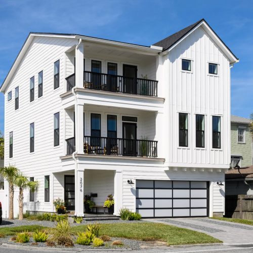 A spacious white coastal-style home featuring multiple balconies, large windows, and a two-car garage, set against a bright blue sky with palm trees in the background.