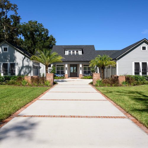 A spacious, contemporary white house featuring a welcoming front porch, surrounded by lush green lawns, palm trees, and well-maintained landscaping under a clear blue sky.