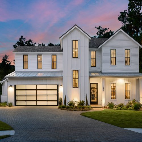 A spacious modern farmhouse with white exterior siding, large windows, and an attached two-car garage, set against a scenic sunset sky with lush landscaping.