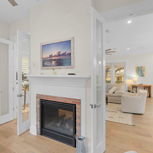 A bright living room featuring a cozy fireplace with a white mantel, surrounded by large windows with white shutters, and an open layout leading to a dining area and additional seating.