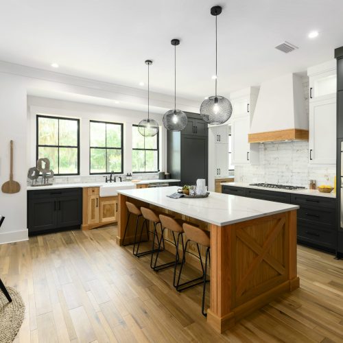 Bright, modern kitchen featuring black and wood cabinetry, a large island with seating, and stainless steel appliances, illuminated by natural light from large windows.