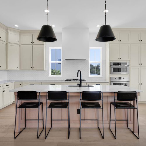 Modern white kitchen featuring a large island with a wooden countertop, black bar stools, and black pendant lighting. The space includes built-in appliances and large windows providing natural light.