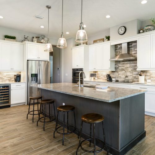 Spacious kitchen featuring white cabinetry, a large island with seating, stainless steel refrigerator, oven, and range hood, illuminated by pendant lights.