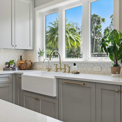 Bright kitchen featuring a large window with a view of lush trees and palm trees, white farmhouse sink, and light gray cabinetry with gold hardware.