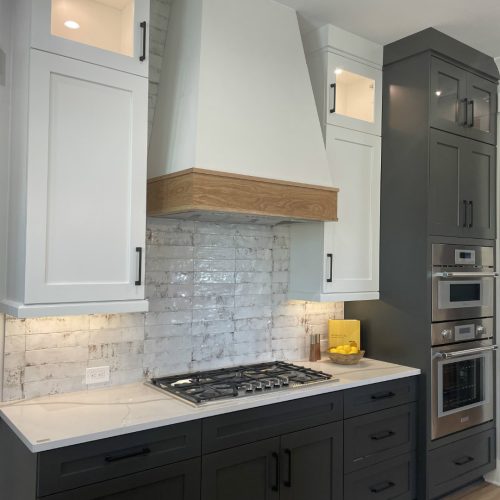 Modern kitchen with black lower cabinets, white upper cabinets, a white brick backsplash, and built-in stainless steel appliances, featuring a central range hood with wooden trim.