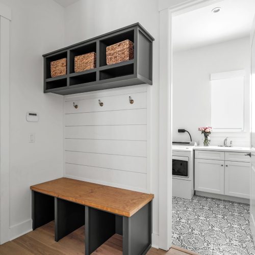 An image of a clean, modern mudroom featuring a wooden storage bench, black cubbies with woven baskets, and hooks for hanging coats, leading into a bright kitchen area.