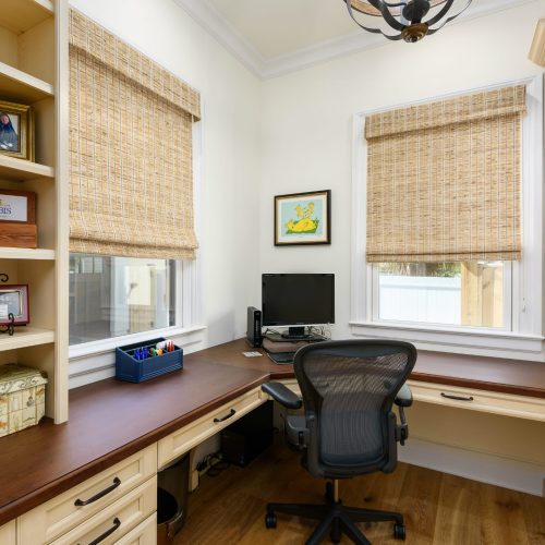An organized home office featuring built-in shelves, a large wooden desk, and a comfortable ergonomic chair, illuminated by natural light from two windows with woven shades.