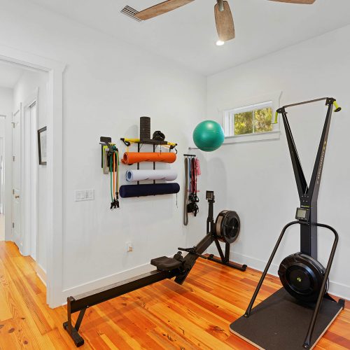 Home gym with various exercise equipment including a rowing machine, stability ball, resistance bands, and a wall-mounted storage rack in a well-lit room.
