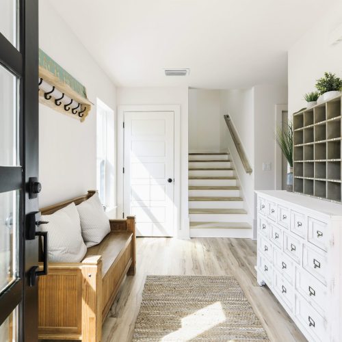 Bright and welcoming entryway featuring a white dresser with multiple drawers, decorative plants, and a wooden bench with cushions. Natural light illuminates the space, creating a cozy atmosphere.