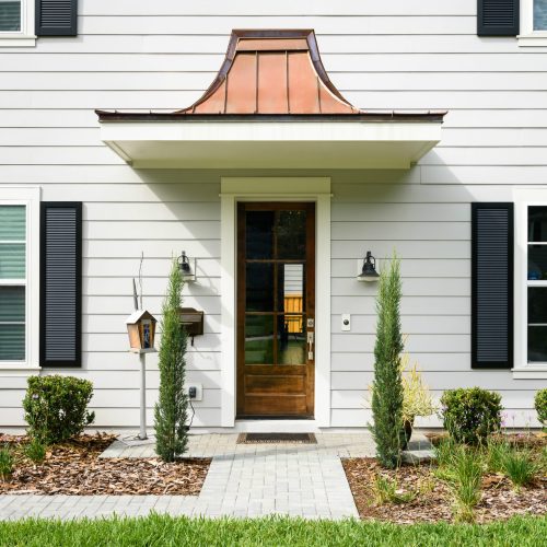 Beautiful white house exterior with black shutters, a wooden front door, and a copper awning, surrounded by well-maintained landscaping and a paved walkway.