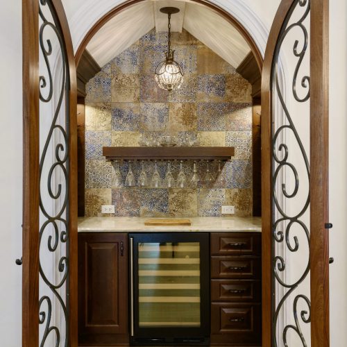 A stylish kitchen wet bar featuring a wine cooler, decorative tile backsplash, and wooden cabinetry, framed by elegant arched double doors with wrought iron details.