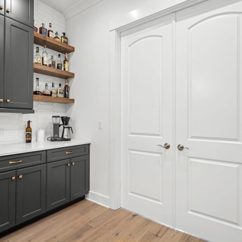 A stylish home bar area featuring dark gray cabinets, white countertops, and open wooden shelves displaying bottles and glassware.