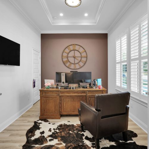 Spacious home office featuring a wooden desk, a comfortable leather chair, and large windows with white shutters, creating a bright and inviting workspace.