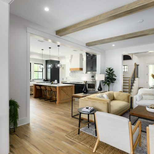 A spacious, bright living room with white walls, wooden beams, and large windows, seamlessly connected to a modern kitchen with an island and pendant lighting.