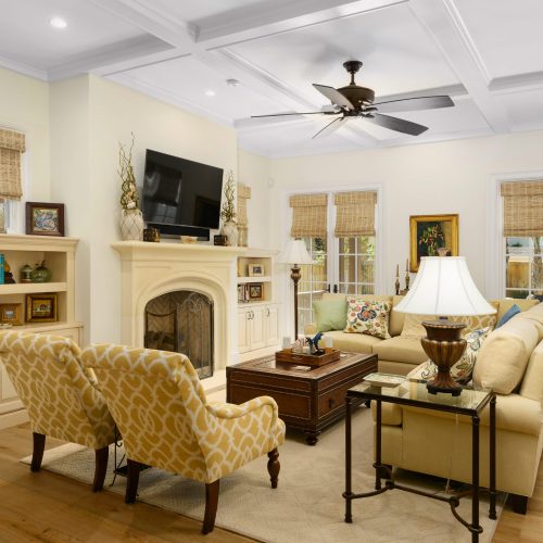Bright and inviting living room with cream-colored sofas, yellow patterned armchairs, and a cozy fireplace, featuring large windows with woven shades and a ceiling fan.