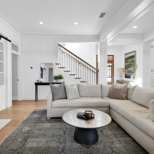 Bright and spacious living room featuring a large L-shaped sofa, a round coffee table, and a fireplace, with natural light streaming through windows and a staircase leading upstairs.