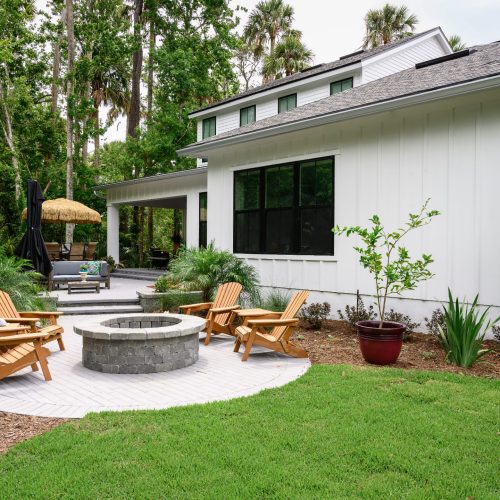 A spacious backyard featuring a modern white house with large windows, a cozy outdoor seating area with Adirondack chairs around a fire pit, surrounded by lush greenery and tall trees.