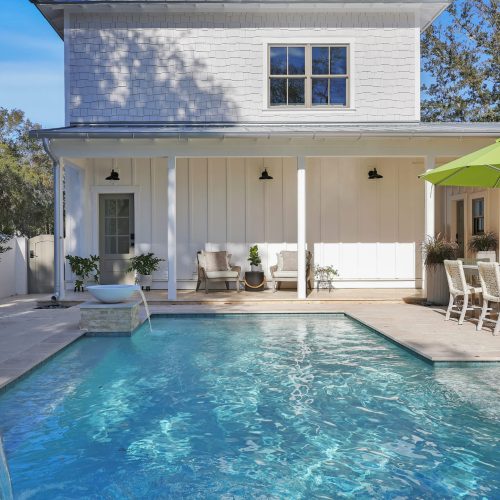A backyard featuring a swimming pool with clear blue water, adjacent to a white house with a covered patio, outdoor dining table with chairs, and a large green umbrella for shade.