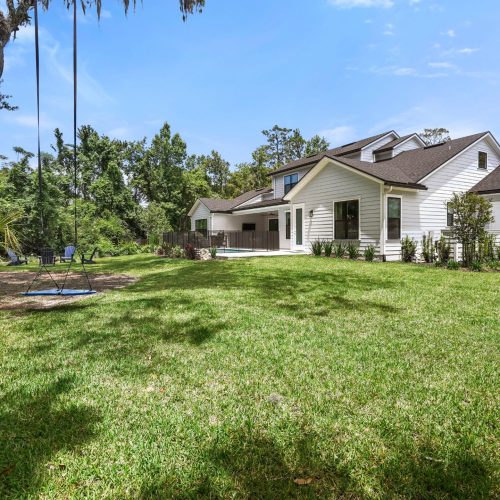 A white, modern-style house with a large, well-maintained backyard featuring lush green grass, mature trees, and a clear blue sky, showcasing a peaceful residential setting.