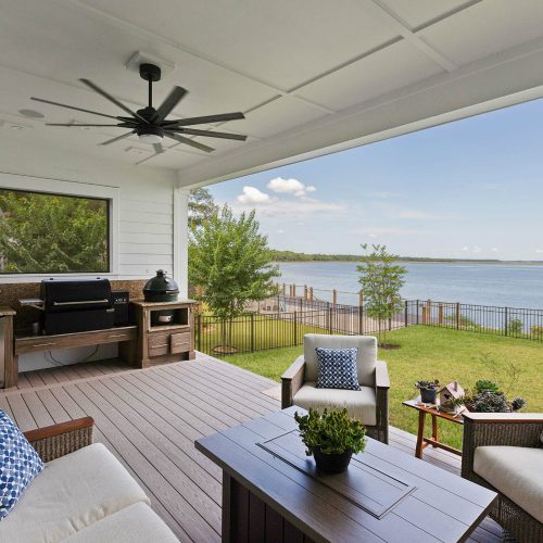 A spacious covered porch overlooking a serene lake with outdoor seating, a ceiling fan, and a view of the water and sky, perfect for relaxing and enjoying nature.