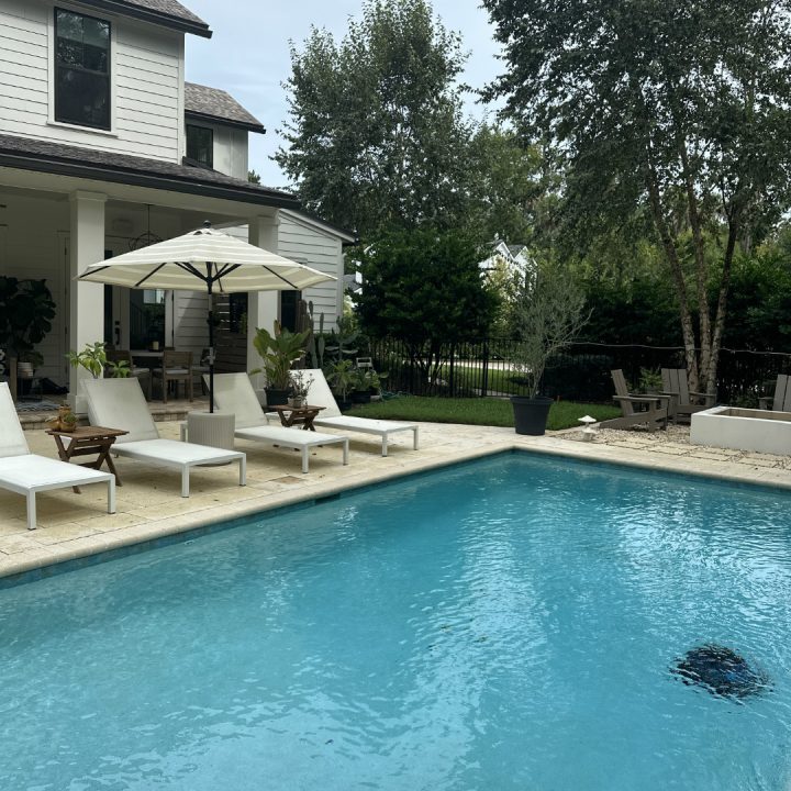 A luxurious backyard with a swimming pool, white lounge chairs, an umbrella, and lush greenery, showcasing modern outdoor living space designed by John Merrill Homes.