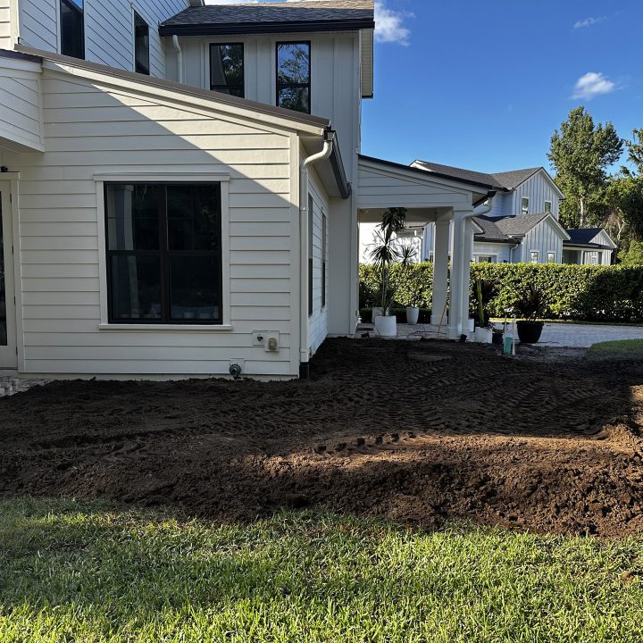 Newly landscaped backyard under construction at a modern white house with lush green grass, potted plants, and a clear blue sky, showcasing custom home exterior features and garden preparation.
