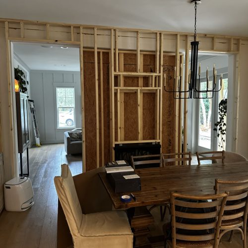 1. An interior view of a dining area under renovation with exposed wooden framing, a black chandelier, and view into the living room with large windows.