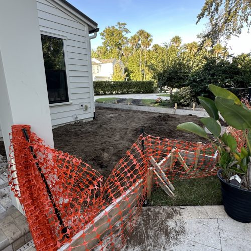 1. Corner of a house with outdoor construction site, dirt ground, and orange safety fencing, with lush green trees and clear blue sky in the background.