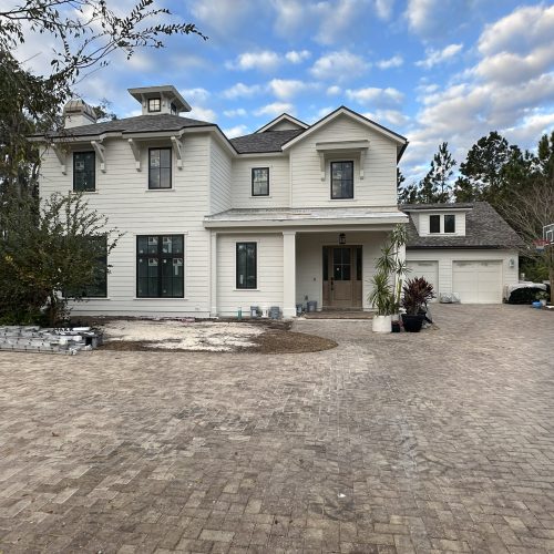 Modern white two-story home with a spacious driveway, surrounded by trees and greenery, under a partly cloudy sky.