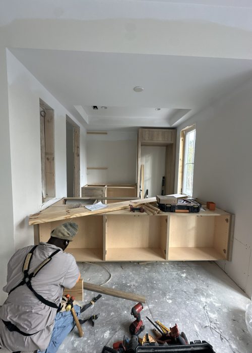 1. Interior view showing kitchen cabinetry under construction with a worker installing custom wooden cabinets in a new home.