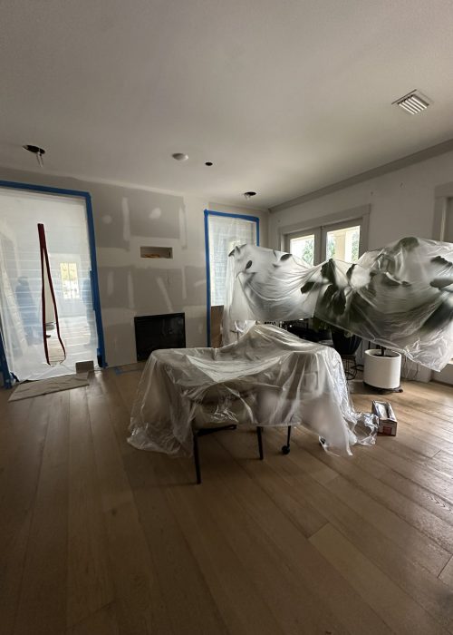 Bright living room under renovation, featuring large windows, hardwood flooring, and a fireplace, with furniture covered in protective plastic. Construction tools and supplies are present as work continues.