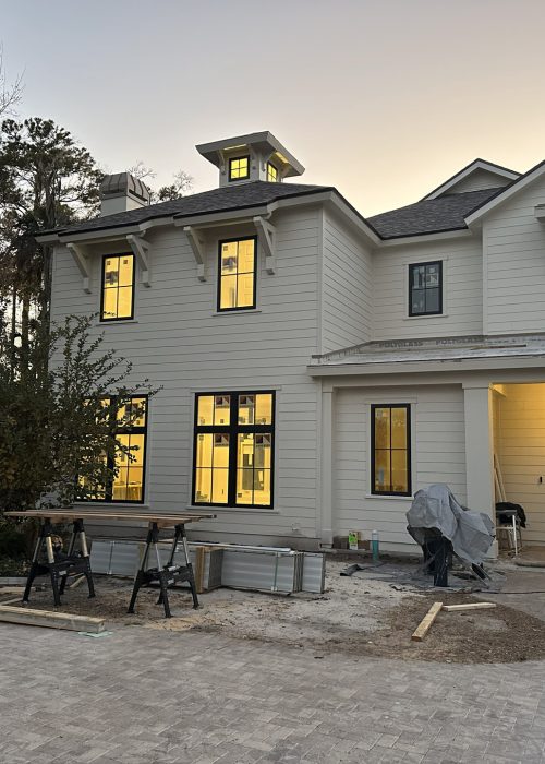 Newly constructed modern white house with warm interior lighting visible through windows, featuring a front porch, multiple stories, and ongoing landscaping or construction work in a peaceful suburban setting.